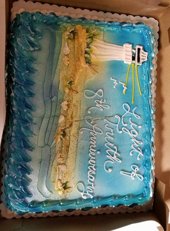 A decorated sheet cake reading Light of Truth Anniversary, iced with a lighthouse and shoreline.