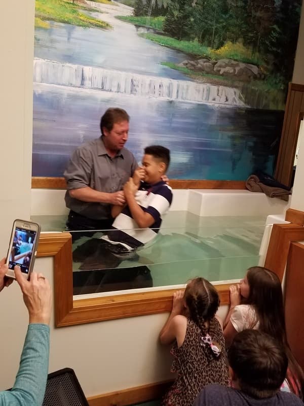 A child being baptized in the LOTC baptistry while family members photograph from the pews.