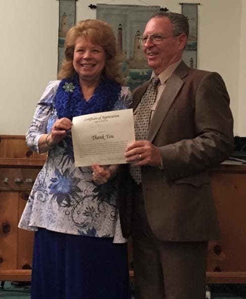 A woman and a man holding a Certificate of Appreciation from Light of Truth Church, standing in front of the sanctuary's lighthouse banners.