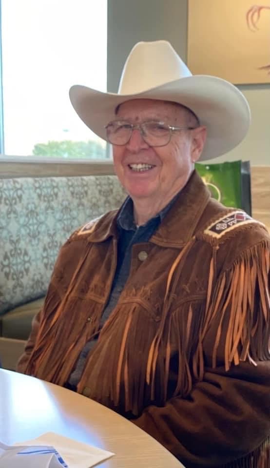 Elder Jerry Benson smiling, wearing a pale cowboy hat and a fringed suede jacket.