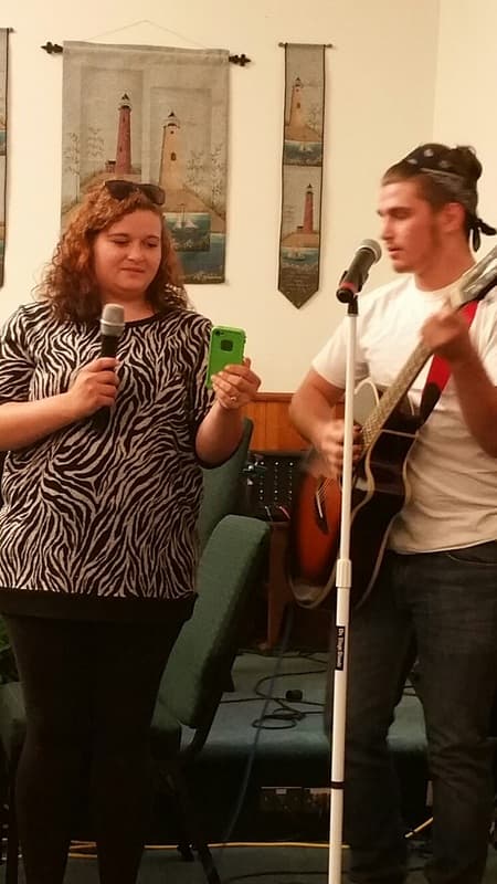 A young woman singing into a microphone beside a guitarist, with lighthouse banners on the sanctuary wall.